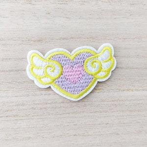 Peut inclure: Un cœur violet avec un contour jaune et des ailes blanches. Le cœur a un plus petit cœur rose au centre.