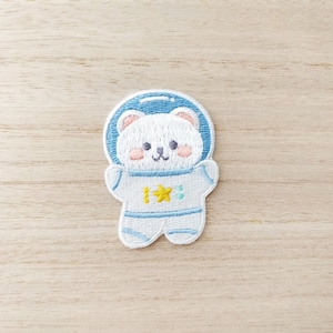 May include: Embroidered patch of a white bear in a blue astronaut suit and helmet. The bear has pink cheeks and a smiling face. The astronaut suit has a yellow star and a turquoise accent. The patch is on a light wood grain background.