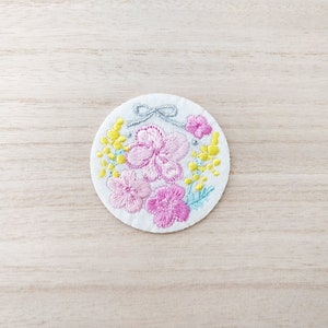 May include: A circular, white embroidered patch with a floral design. The patch features pink flowers, yellow mimosa, and a silver bow. The embroidery is detailed, with the flowers in full bloom. The patch is likely for clothing or accessories.