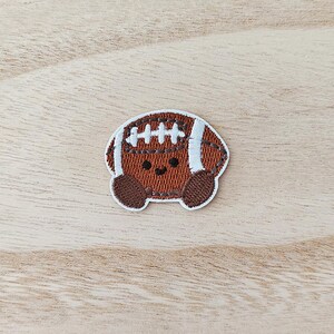 Peut inclure: Écusson brodé d'un ballon de football américain de dessin animé avec un visage souriant. Le patch est marron avec des rayures blanches et des détails de couture. Le fond est un grain de bois clair.