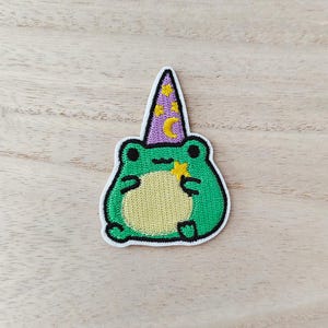 May include: Embroidered patch featuring a green frog wearing a purple wizard hat adorned with yellow stars and a crescent moon. The frog has a yellow belly and a star-shaped wand. The patch is outlined in black and white.