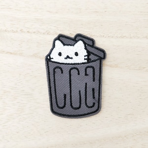May include: Embroidered patch showing a white cat in a grey bin. The bin has the letters "CCG" embroidered on it. The patch is on a light wood grain surface.