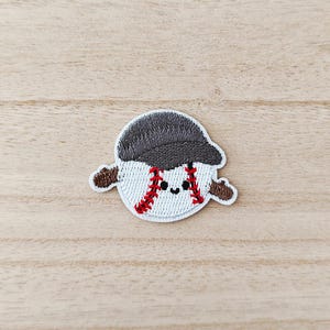 May include: Embroidered patch of a baseball character with a smiling face. The baseball has red stitching and is wearing a grey baseball cap. The patch has brown arms and is on a light wood grain surface.
