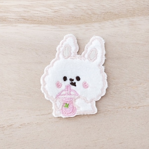 May include: White embroidered patch of a fluffy bunny holding a pink boba tea with a green tapioca pearl.