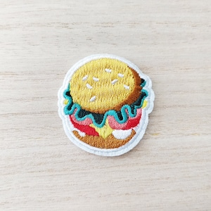 May include: Embroidered iron-on patch of a cartoon burger with a yellow bun, green lettuce, pink tomato, and brown meat.