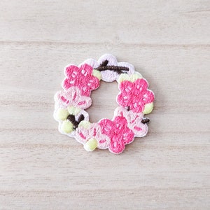 May include: Embroidered floral wreath patch with pink blossoms, light green accents, and brown stems. The circular patch has a detailed, textured appearance, ideal for embellishing clothing or accessories.