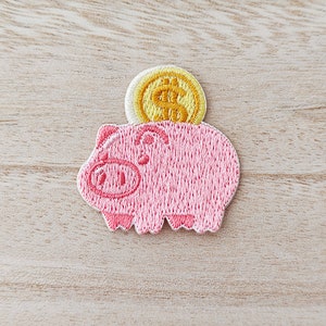 May include: Embroidered patch of a pink piggy bank with a gold coin. The coin features a dollar symbol. The patch is designed for application on fabric items such as clothing or bags. The patch is approximately 5 cm in height.