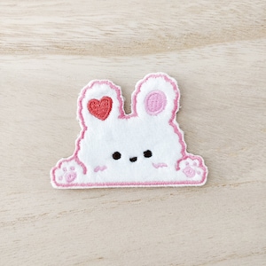 May include: White felt bunny applique with pink embroidered details. The bunny has a heart on one ear and a circle on the other. The bunny is smiling with two black eyes and a pink nose.