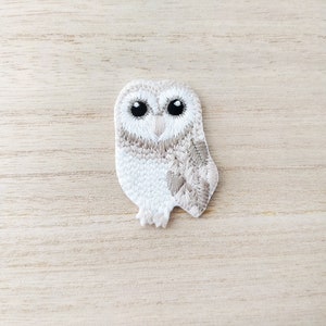May include: White and brown embroidered owl applique patch. The owl has black eyes and a white beak.