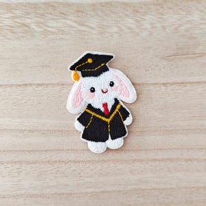 May include: Embroidered patch of a white bunny in a black graduation gown and mortarboard. The bunny has pink ears and a red tie. The patch is intricately stitched, depicting a smiling bunny, perfect for celebrating graduation.