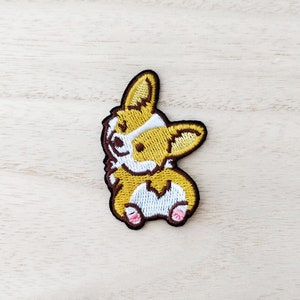 May include: Embroidered patch of a brown and yellow corgi dog with white markings. The corgi is standing on its hind legs with its front paws raised.