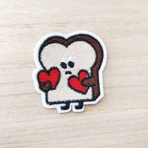 May include: Embroidered patch of a cartoon bread slice with a brown crust, holding two red hearts. The bread has a white face with two black eyes and a small mouth.