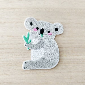 May include: Embroidered iron-on patch featuring a gray koala holding a green eucalyptus leaf. The koala has pink cheeks and black eyes.