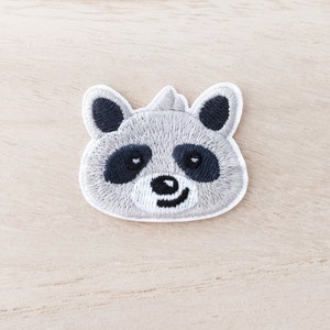 Peut inclure: Écusson brodé représentant un visage de raton laveur souriant. Le patch a un corps gris, des yeux et des accents d'oreilles noirs, et une bordure blanche. Le visage du raton laveur est détaillé avec des marques noires autour des yeux et un petit sourire incurvé.