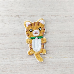 May include: Embroidered iron-on patch of a cute orange tabby cat wearing a green collar. The cat has white fur on its chest and belly, and a white tail.