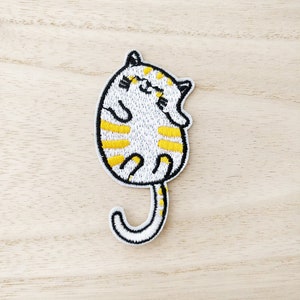 May include: White and yellow embroidered iron-on patch of a sleeping cat with a curled tail.