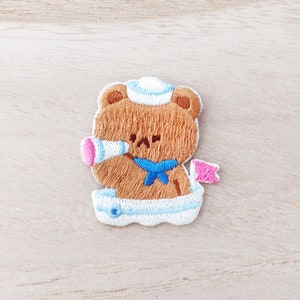 May include: Embroidered brown bear wearing a blue and white sailor hat, holding a pink megaphone and a pink flag. The bear is sitting in a white boat with blue trim.