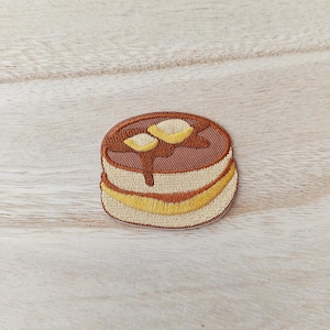 May include: An embroidered patch shaped like a stack of two pancakes. The top pancake is brown with two yellow butter pats and a brown syrup drizzle. The bottom pancake is light tan with a yellow horizontal stripe representing a layer of filling.