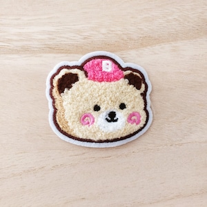 Peut inclure: Un ours en peluche brun et blanc avec un chapeau rose sur lequel est brodée une lettre "B" blanche. L'ours a des joues roses et un nez blanc.