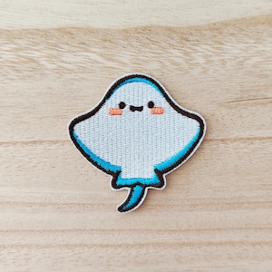 Peut inclure: Un écusson brodé bleu clair en forme de raie manta. L'écusson a un contour noir avec une bordure bleu turquoise. La raie manta a un visage souriant avec des joues couleur pêche. L'écusson est sur une surface en bois brun clair.