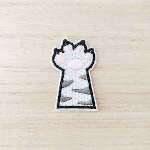 May include: A white and gray cat paw embroidered patch with pink paw pads. The patch is outlined in black thread.