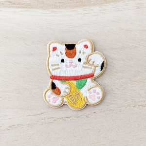 May include: Embroidered patch of a white Maneki-Neko, a Japanese lucky cat, with orange and black markings. The cat is holding a gold coin with the Japanese word "lucky" written on it.