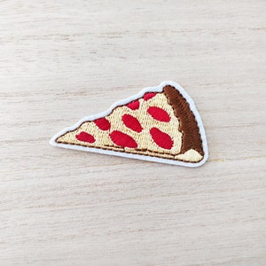 May include: Embroidered patch in the shape of a pizza slice. The patch features a white border, a tan crust, and a brown edge. Red pepperoni dots are scattered across the tan pizza base. This patch is perfect for adding to clothing or accessories.