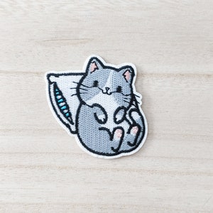 May include: Embroidered iron-on patch of a grey cat curled up with a white pillow. The cat has a white face and pink paws.