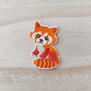 May include: Embroidered patch of a cartoon red panda in orange, red, and white. The patch shows the red panda sitting with its paws crossed. The patch is on a light wood grain surface.