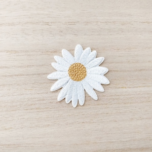 Peut inclure: Patch brodé de couleur blanche représentant une marguerite avec un centre jaune. Le patch est posé sur une surface en bois brun clair.