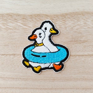 May include: Two white embroidered geese with yellow beaks and feet are floating in a blue inflatable ring. The geese are facing the same direction and the one in the back has a yellow collar.