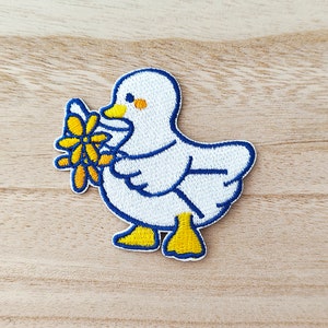 May include: Embroidered patch of a white duck with blue outlines, yellow feet and beak, holding a yellow flower with a blue outline. The patch is designed for sewing or ironing onto fabric.