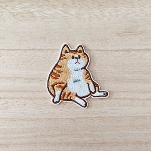 May include: Embroidered iron-on patch of a cartoon orange tabby cat with white markings sitting with its legs crossed. The cat has a white belly and a brown nose.