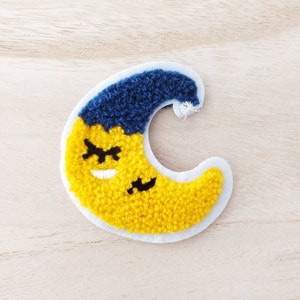 May include: A yellow crescent moon with a blue sleeping cap, embroidered with a smiling face and closed eyes.