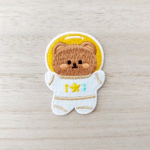 May include: A brown bear wearing a white astronaut suit with a yellow star on the chest. The bear is embroidered on a white fabric.
