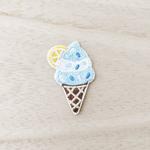 May include: Embroidered patch of a light blue ice cream cone with white and blue details, and a yellow lemon slice. The cone is brown with a waffle pattern. Ideal for adding to clothing or accessories.