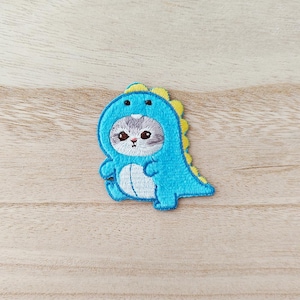 May include: Embroidered patch of a grey and white cat in a blue dinosaur costume with yellow detailing. The cartoon-style patch is ideal for customising clothing or accessories. The patch is approximately 7.6 cm tall.