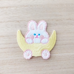 May include: Embroidered patch featuring a white bunny with pink accents, sitting on a yellow crescent moon. The bunny has blue eyes and pink paws. The patch is designed for decorative use on clothing or accessories.
