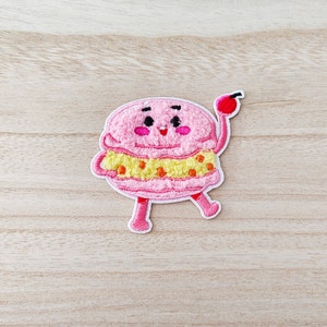 Peut inclure: Un macaron rose avec une bouée de sauvetage jaune et orange, souriant avec des joues roses et une cerise sur le dessus, porte un contour blanc et a deux jambes roses.