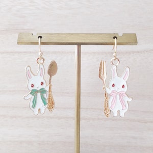 May include: A pair of gold-tone earrings featuring white bunny charms holding a spoon and fork. The bunnies have pink accents and red eyes.