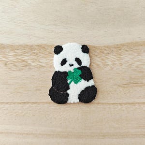 May include: Embroidered panda bear patch in black, white, and green. The panda is holding a green shamrock. The patch is made of textured fabric and is set against a light wood grain background. The panda has black ears, eyes, and limbs.