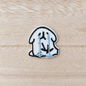May include: Embroidered patch depicting a white cartoon character with black outlines and blue details. The character is shown crying, with blue lines symbolizing tears. The patch has a black border and is on a light wood surface.