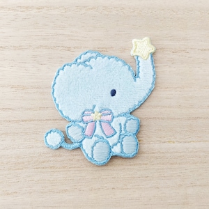 May include: A light blue embroidered elephant holding a yellow star. The elephant is wearing a pink bow with a yellow centre.