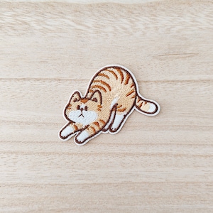 May include: Embroidered iron-on patch of a brown and white tabby cat stretching. The cat has a white patch on its chest and a white stripe on its face.