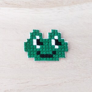 May include: Green pixelated alien face with white eyes and a black mouth, embroidered on a white fabric.