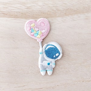 May include: Embroidered patch of an astronaut holding a pink heart-shaped balloon. The astronaut wears a grey spacesuit and a blue helmet. The balloon contains colourful confetti.