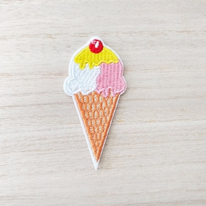 May include: A three-scoop ice cream cone with a red cherry on top. The ice cream is yellow, white, and pink. The cone is brown and has a waffle texture.
