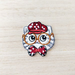 May include: Embroidered iron-on patch of a grey cat wearing red and white polka dot glasses, a red and white polka dot hat, and a red and white polka dot bow tie.