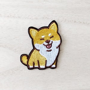 May include: Embroidered patch of a smiling Shiba Inu dog. The patch has a yellow and white body with a brown outline. The dog's tongue is pink, and its eyes are closed. Ideal for adding to clothing or accessories.