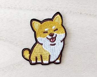 Puede incluir: Parche bordado con un perro Shiba Inu sonriente. El parche tiene un cuerpo amarillo y blanco con un contorno marrón. La lengua del perro es rosa y sus ojos están cerrados. Ideal para añadir a ropa o accesorios.